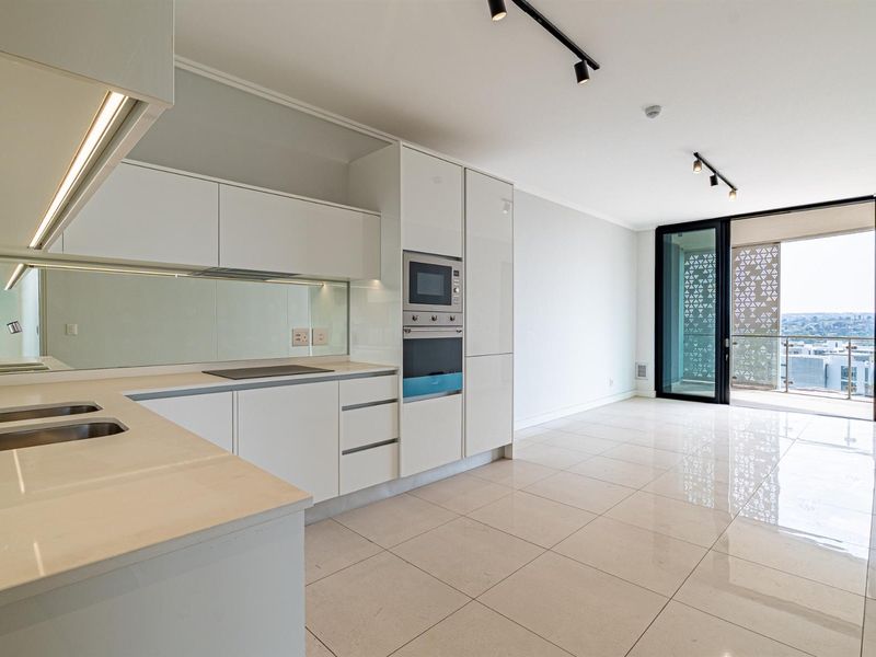 Modern open-plan kitchen and living area featuring sleek white cabinetry, integrated appliances, stone countertops, and large sliding doors leading to a private balcony.