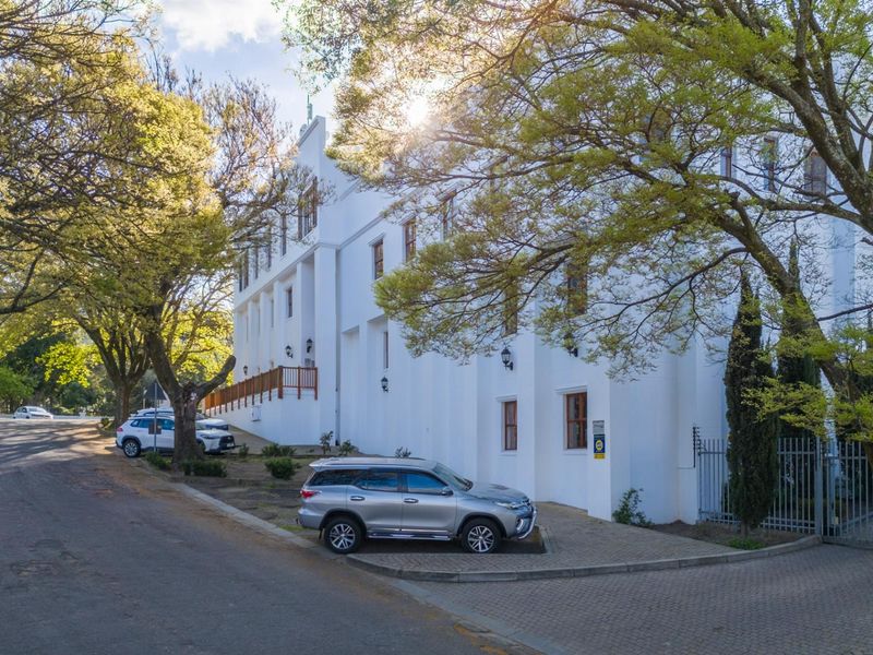 A view of the office building exterior featuring white facade, wooden balcony railing, and paved parking area surrounded by mature trees.
