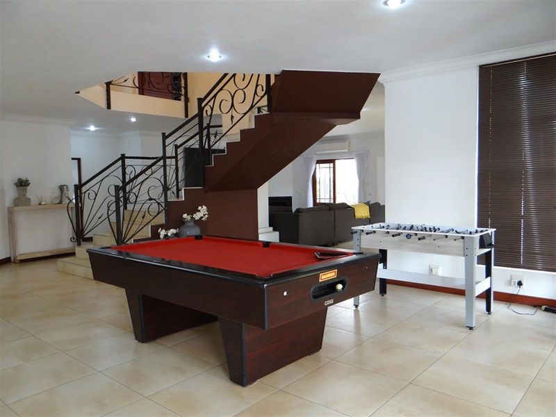 Spacious open-plan living area featuring tiled flooring, a prominent staircase with decorative wrought iron railing, and large windows with venetian blinds.