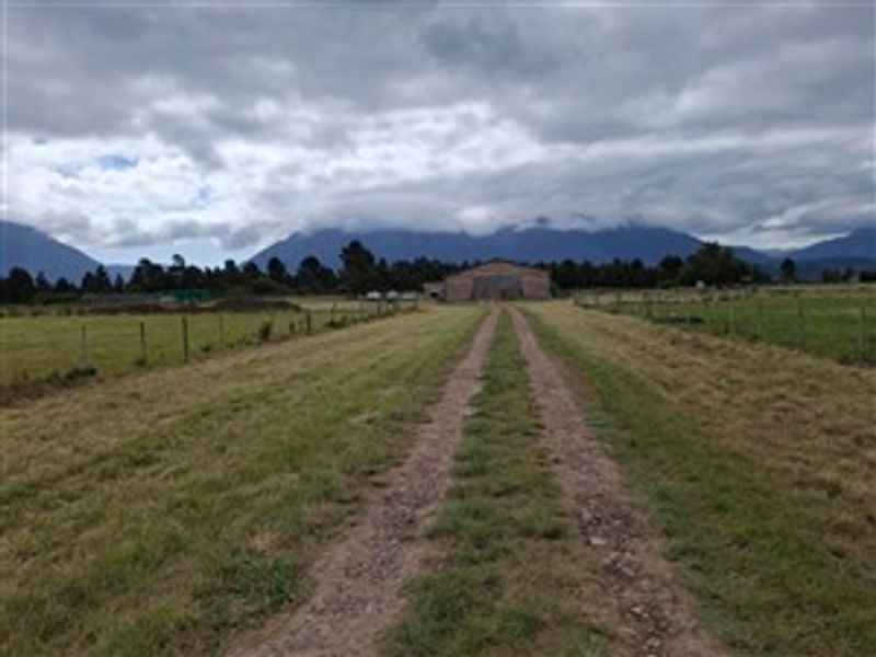A wide-angle view of the farm property featuring a long dirt driveway leading towards a large barn structure set against a mountain backdrop.