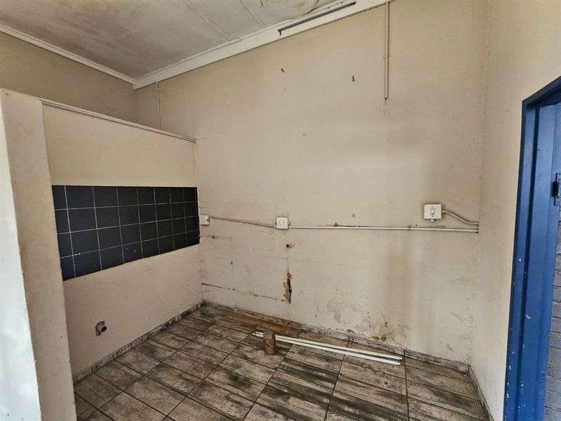 A vacant utility or kitchen prep area featuring tiled flooring, partial wall tiling, and exposed electrical infrastructure, requiring significant refurbishment.
