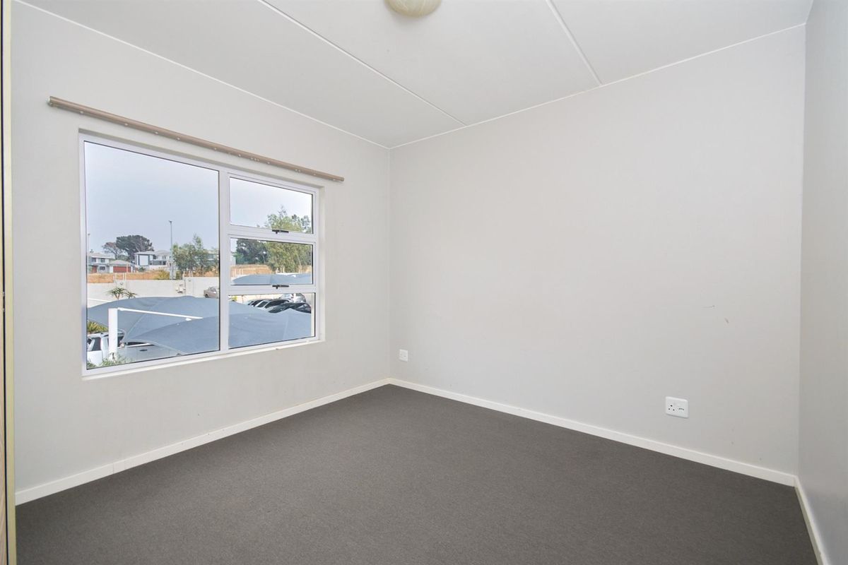 A clean, empty bedroom featuring neutral walls, carpeted flooring, and a large window providing natural light and views of the complex parking area.
