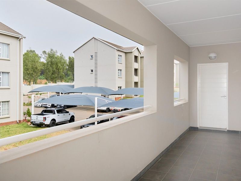 A clean, covered balcony area featuring tiled flooring, a modern balustrade, and an exterior wall light, overlooking the apartment complex parking area.