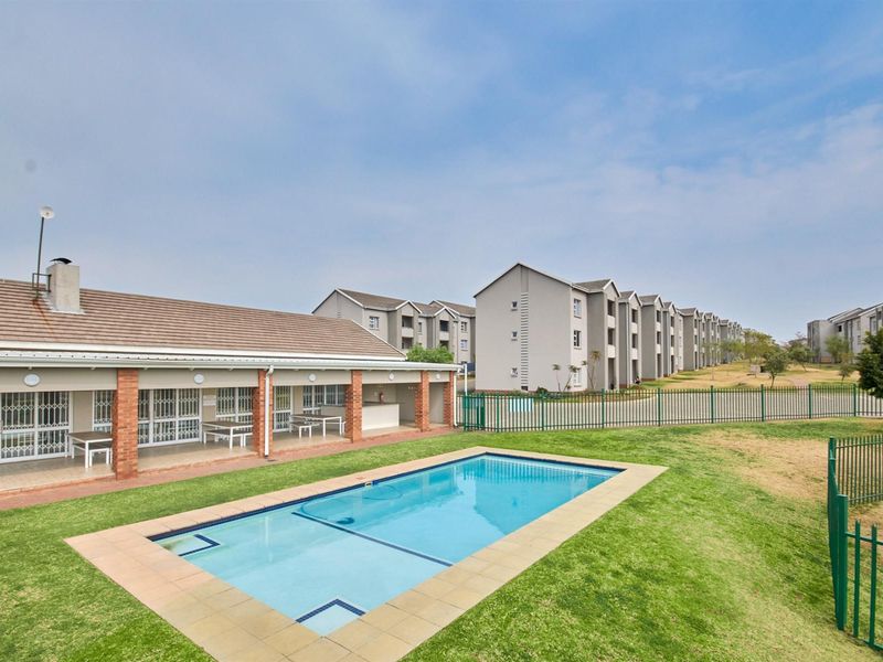 Communal swimming pool area featuring a paved surround, adjacent brick clubhouse with covered patio, and well-maintained lawn within a secure apartment complex.