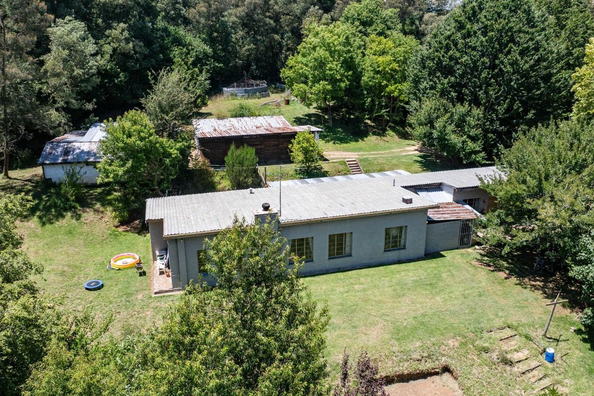 Aerial view of a residential property featuring a main house with a corrugated metal roof, surrounded by expansive garden grounds and secondary outbuildings.