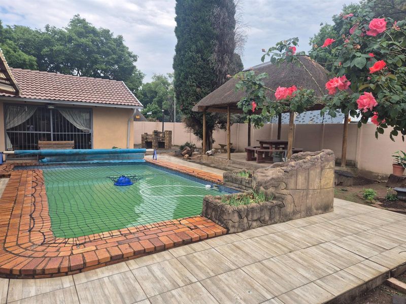 Outdoor entertainment area featuring a swimming pool with safety net, a thatched lapa, and a built-in brick braai set within a paved garden space.