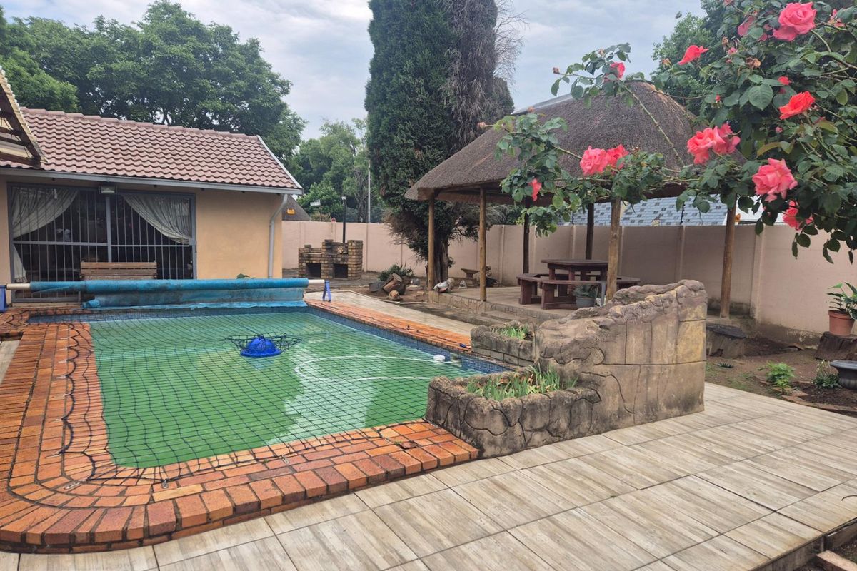 Outdoor entertainment area featuring a swimming pool with safety net, a thatched lapa, and a built-in brick braai set within a paved garden space.
