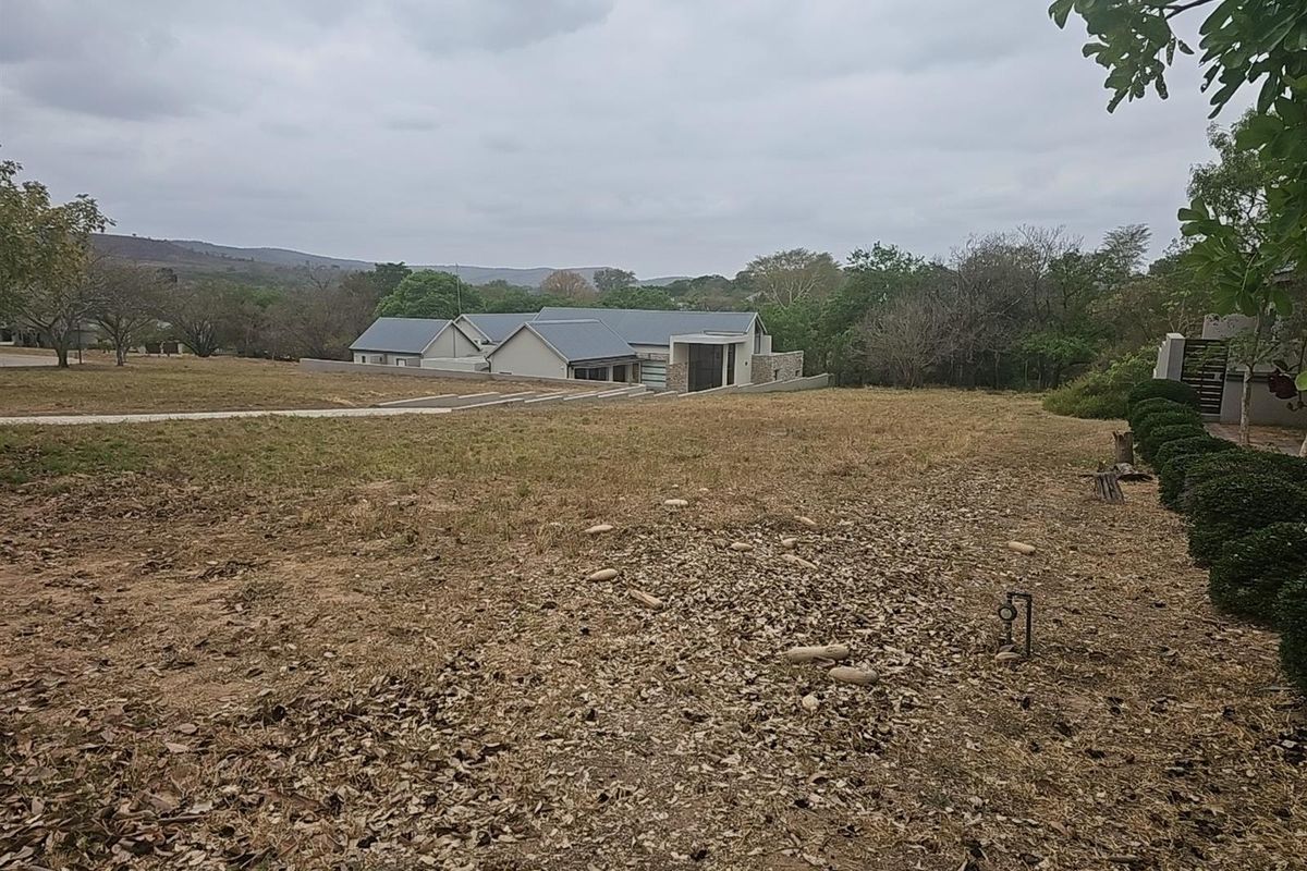 A view of a vacant residential plot featuring natural landscaping, dry grass, and a distant view of a modern house structure.