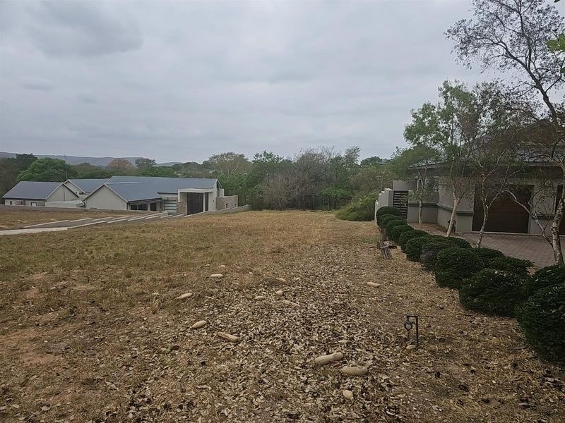 A view of a vacant residential plot featuring established landscaping, adjacent modern housing, and a paved driveway area.