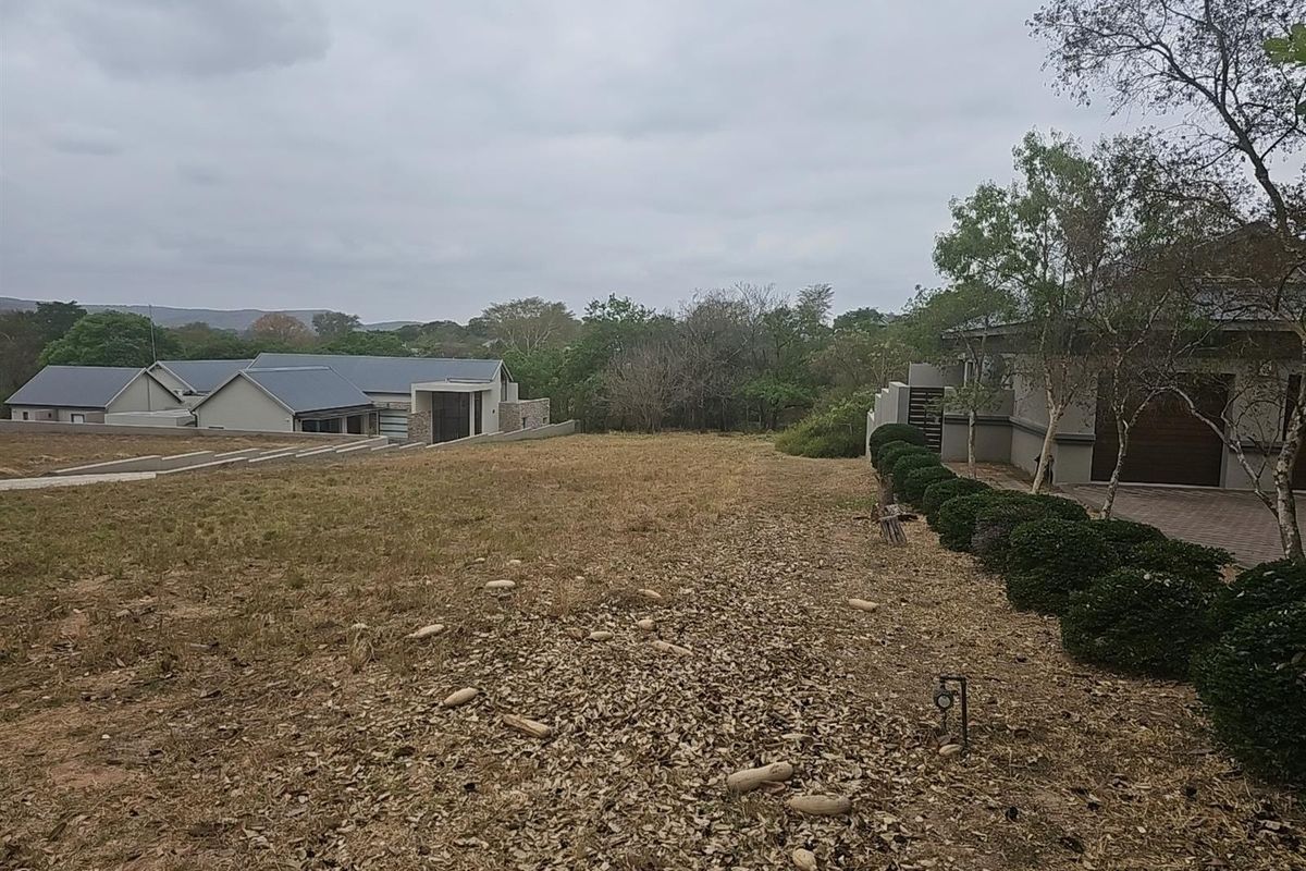 A view of a vacant residential plot featuring established landscaping, adjacent modern housing, and a paved driveway area.