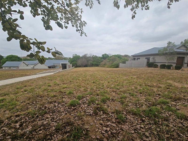 A view of a vacant residential plot featuring natural grass coverage, surrounded by established neighboring properties and mature trees.