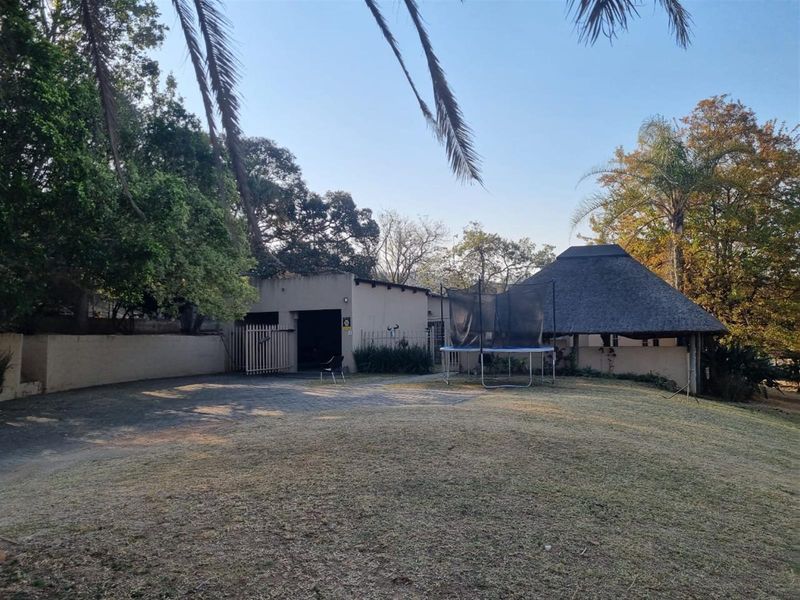 Rear garden area featuring a paved driveway, a traditional thatch roof lapa, and established trees, providing a spacious outdoor entertainment setting.