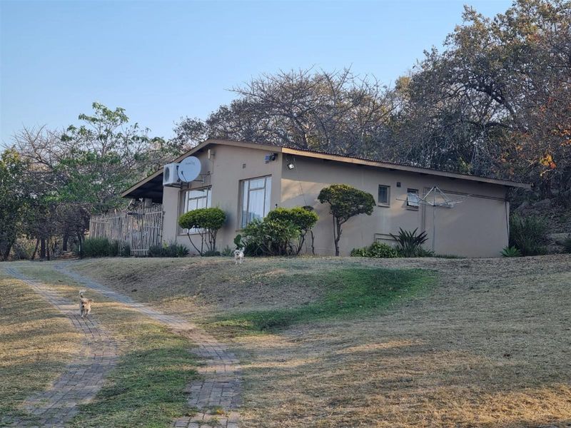 Rear view of the property featuring a paved driveway, exterior wall-mounted air conditioning unit, and security fencing set against a natural landscape.
