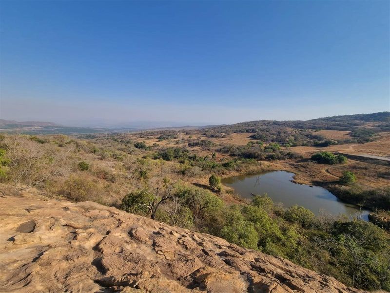 A scenic elevated view overlooking the natural landscape, featuring rolling hills, indigenous vegetation, and a small dam.