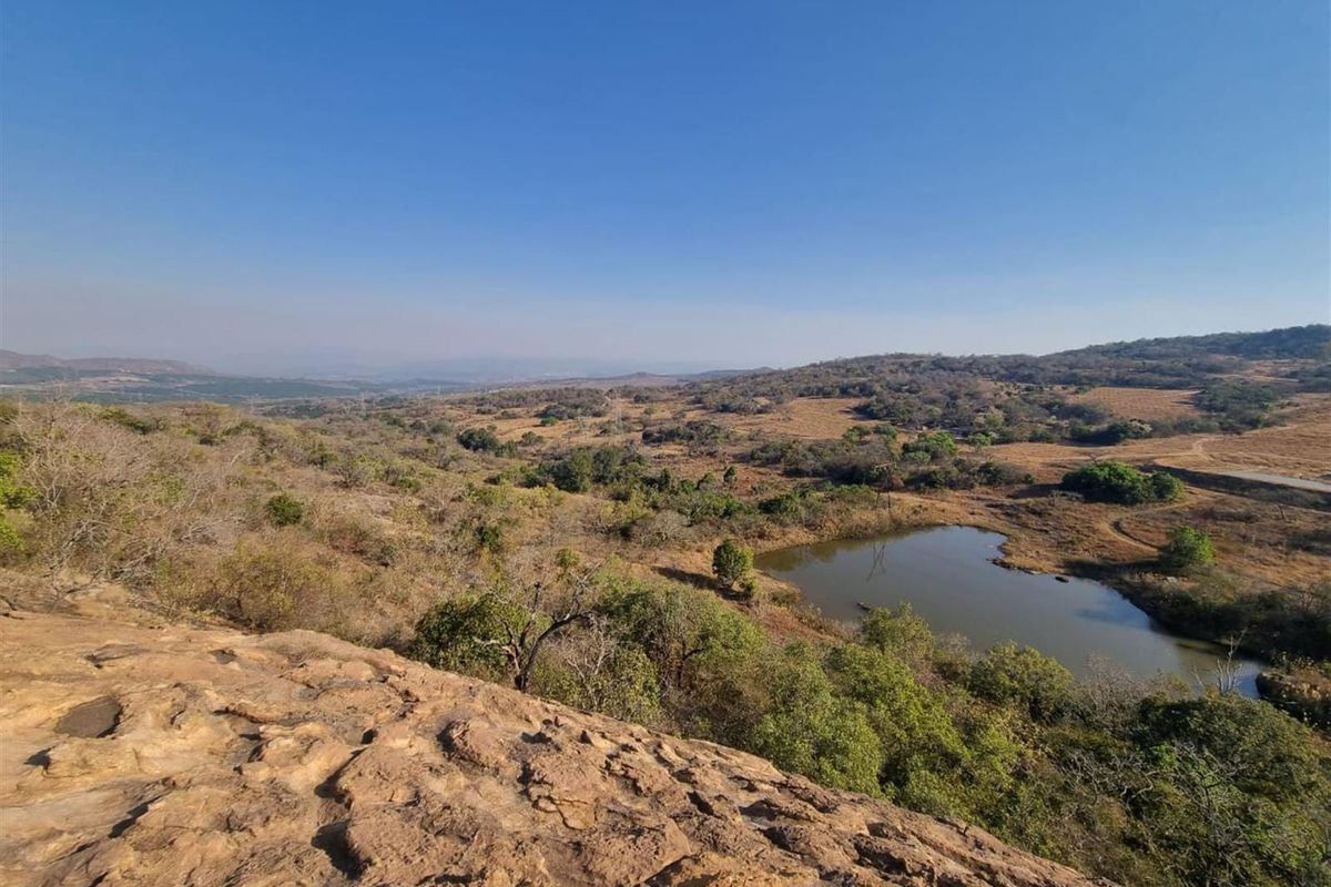 A scenic elevated view overlooking the natural landscape, featuring rolling hills, indigenous vegetation, and a small dam.