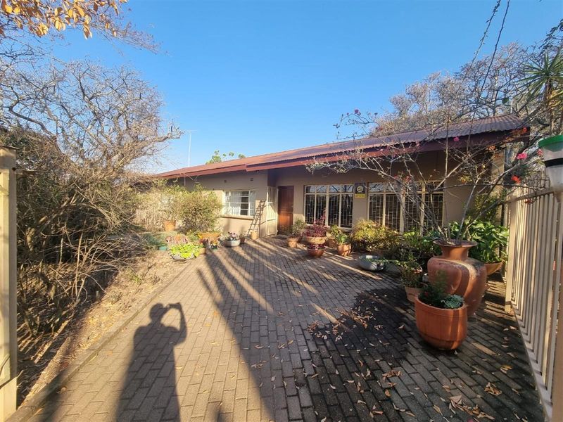 Front exterior view of a single-story house featuring a paved driveway, brick facade, and metal roofing with security bars on the windows.