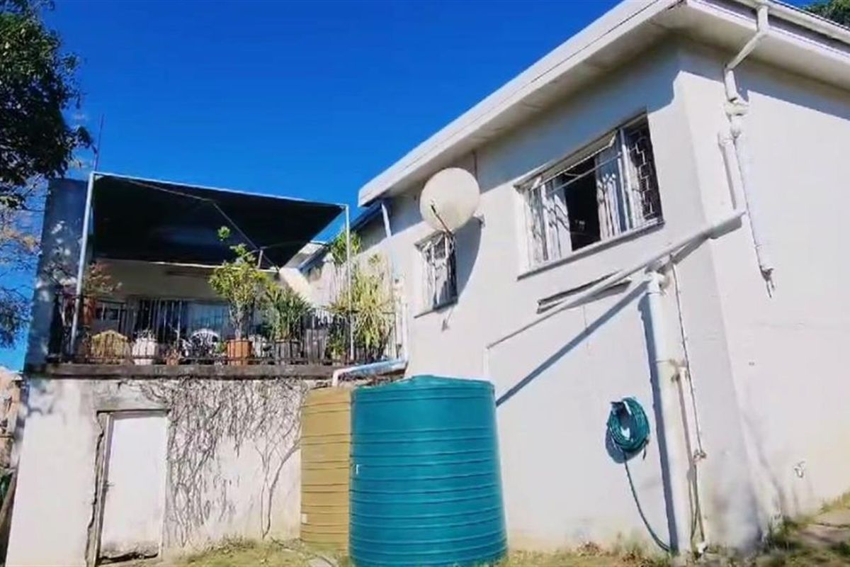 Rear view of the property featuring a raised patio area, water storage tanks, and exterior wall finishes requiring maintenance.