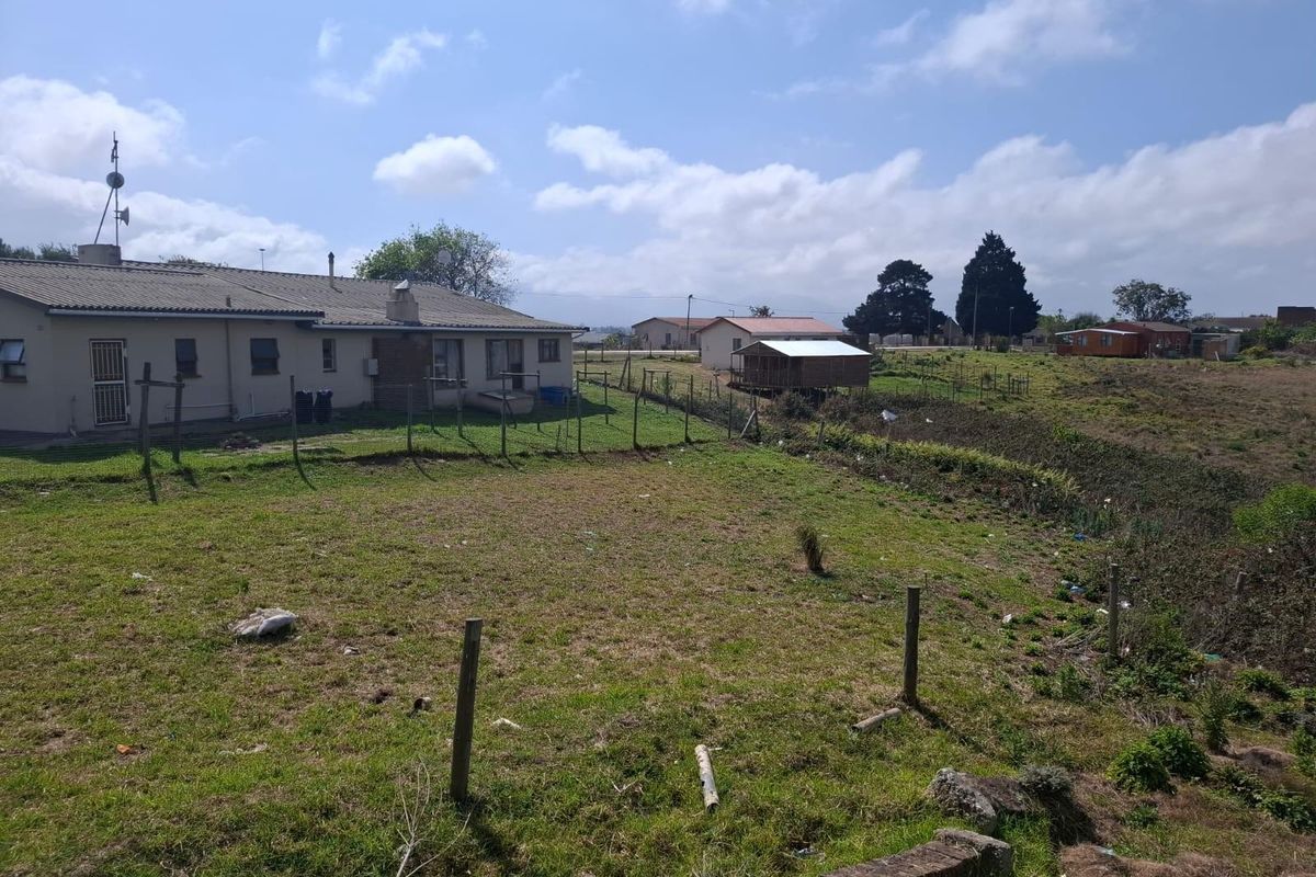 A view of a vacant commercial land plot featuring grass terrain, perimeter wire fencing, and adjacent residential structures in the background.