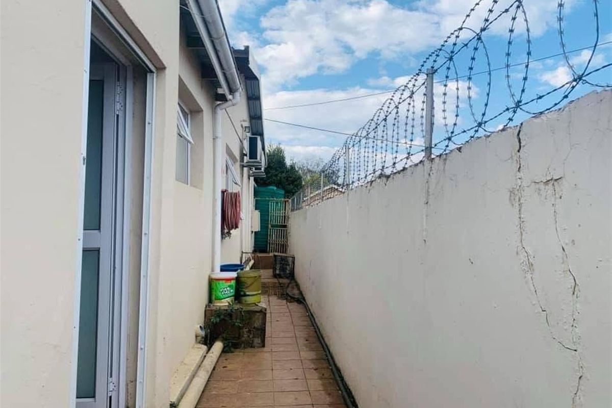 A narrow exterior side passage featuring a tiled walkway, perimeter wall with razor wire, and exterior building access.
