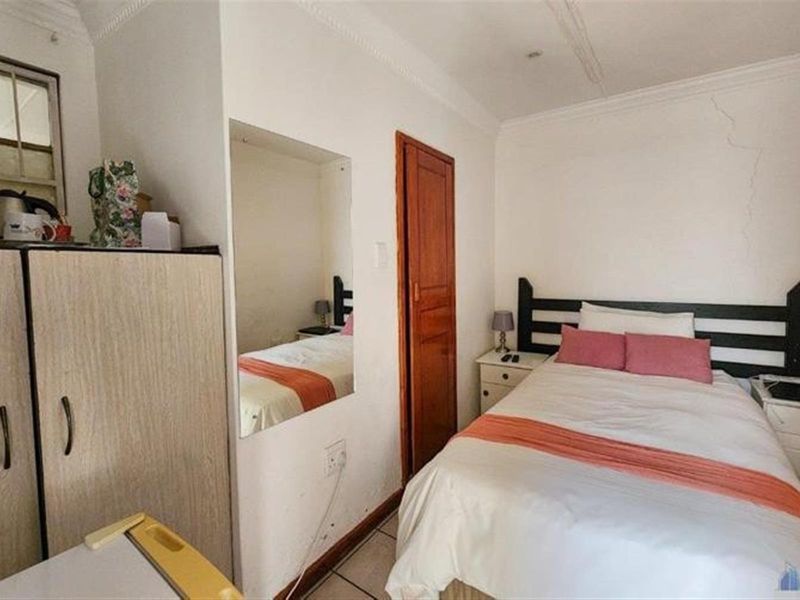 A compact bedroom featuring tiled flooring, a built-in mirror, and a wooden door, showing signs of wall cracking requiring maintenance.