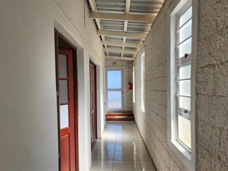 A long, tiled hallway featuring exposed ceiling beams, multiple timber doors, and louvre windows along a textured masonry wall.