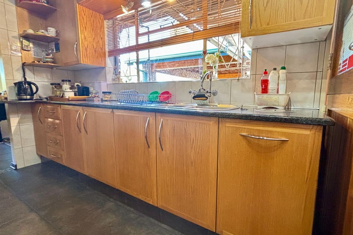 A functional kitchen featuring oak veneer cabinetry, dark laminate countertops, a stainless steel sink, and tiled backsplash with neutral floor tiles.