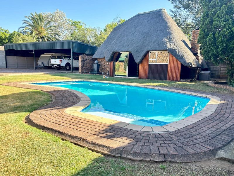 Outdoor pool area featuring a swimming pool with brick paving, adjacent to a unique thatch-roof structure and a spacious carport.