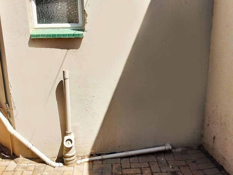Exterior view of a building corner featuring exposed plumbing pipes, a small window with a green sill, and paved ground surface.