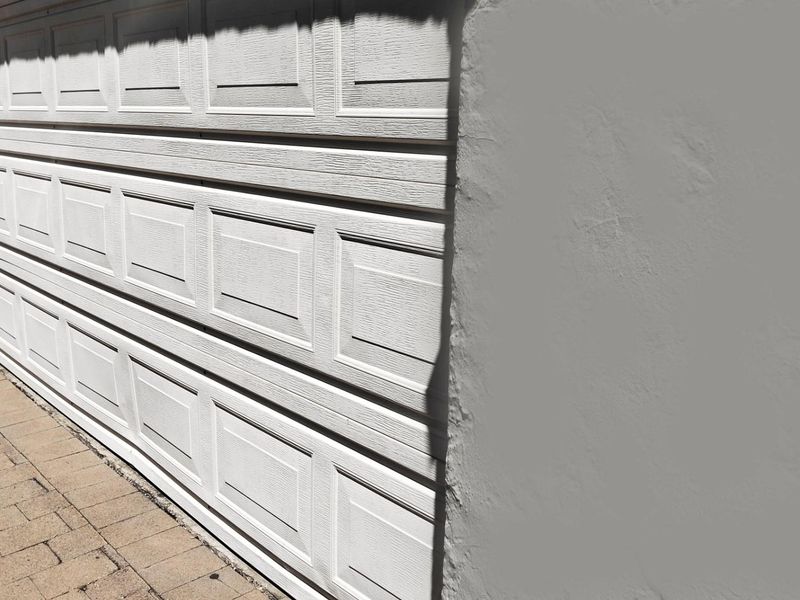 A view of a white sectional garage door with a paved driveway and textured exterior wall.