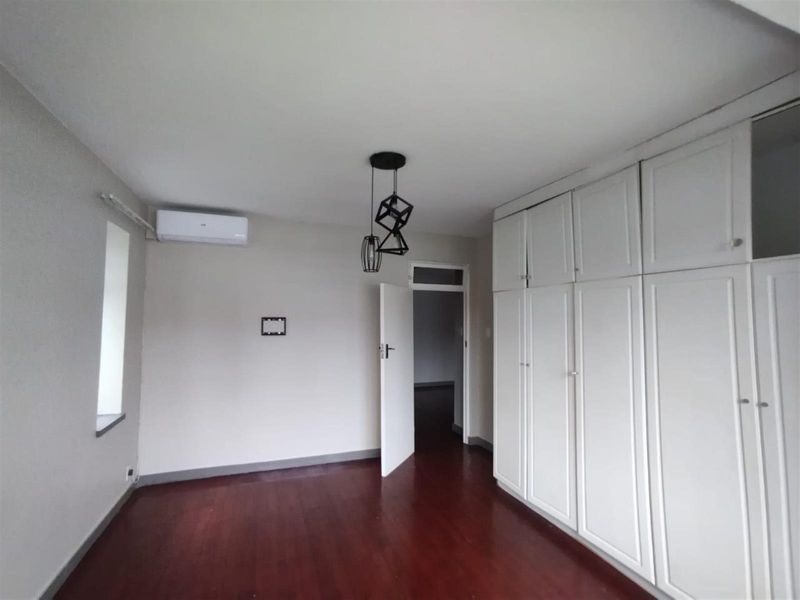A bright, empty bedroom featuring dark laminate flooring, extensive white built-in cupboards, and a wall-mounted air conditioning unit.