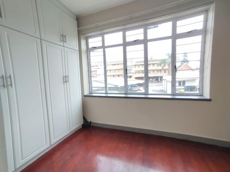 A bright, empty bedroom featuring polished wooden flooring and ample built-in cupboard storage, complemented by a large window with security bars.