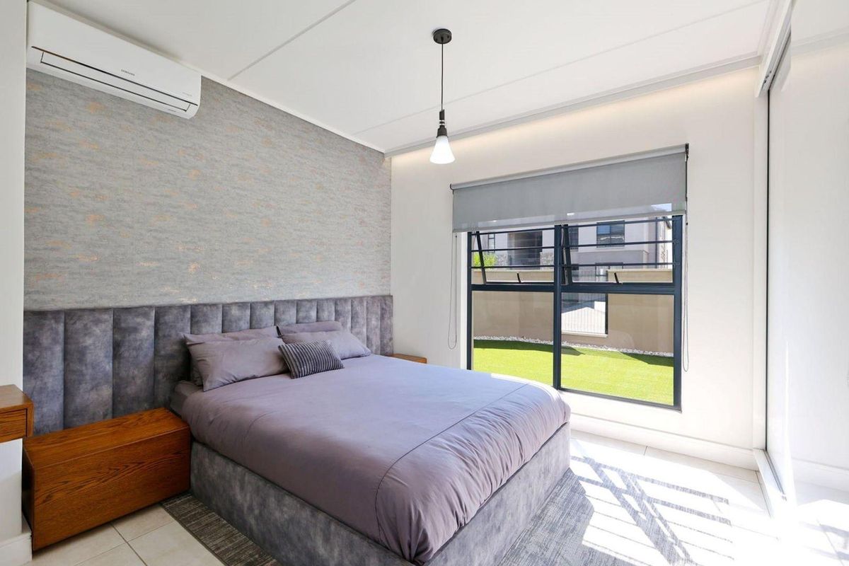 A modern, well-maintained bedroom featuring tiled flooring, a split-system air conditioner, and large windows with roller blinds overlooking a garden area.