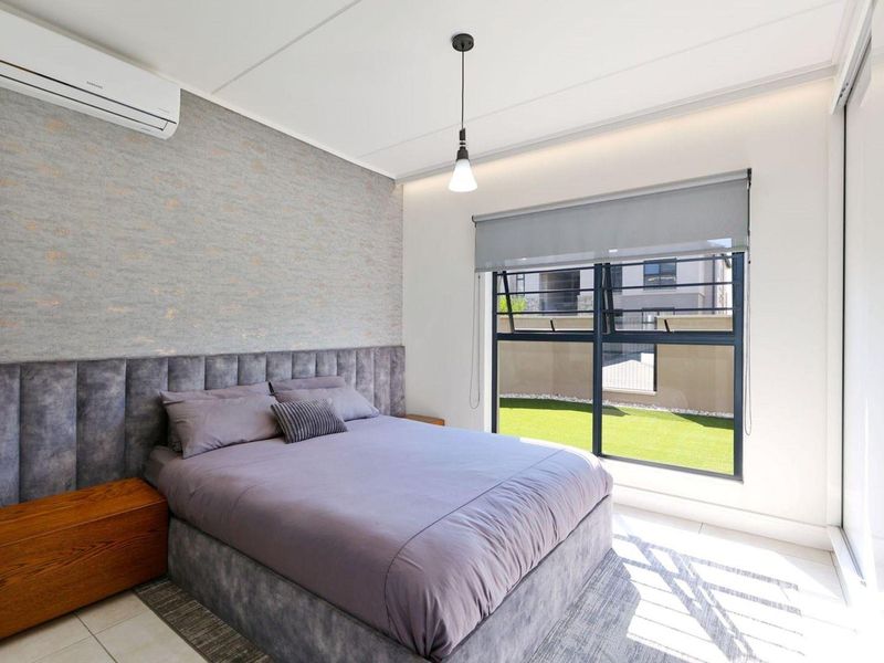 A modern, well-maintained bedroom featuring tiled flooring, a split-system air conditioner, and large windows with roller blinds overlooking a garden area.