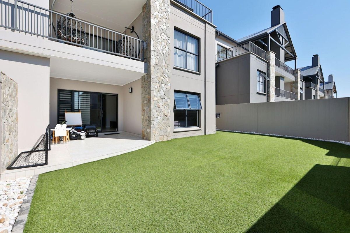 Rear view of the apartment featuring a covered patio, stone-clad pillar, balcony with metal railings, and a well-maintained artificial lawn garden.