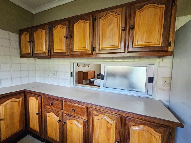 A dated kitchen featuring traditional wooden cabinetry, laminate countertops, and a tiled backsplash with a serving window looking into the adjacent room.