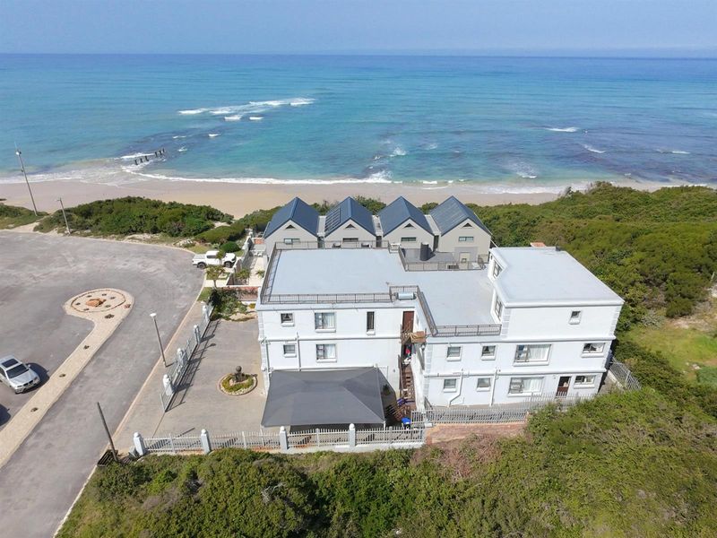 Aerial view of a multi-story coastal property featuring a metal roof, paved driveway, and perimeter fencing, situated directly adjacent to the beach.