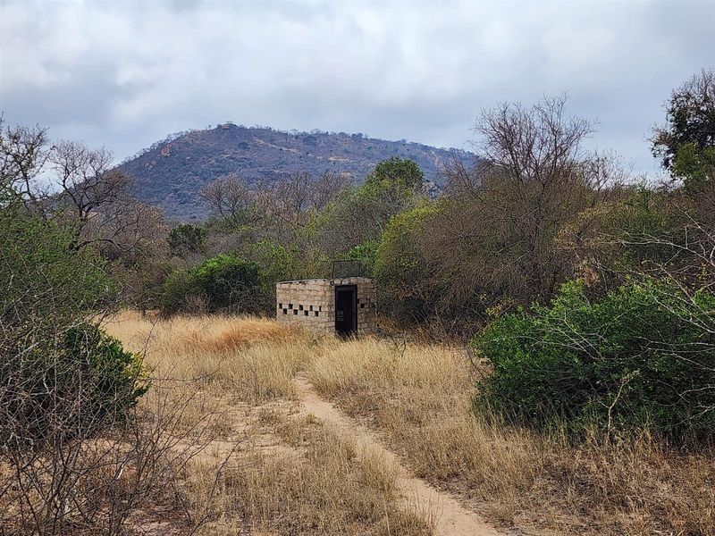 A small, detached stone utility structure situated in a rural landscape, surrounded by natural vegetation and mountain views.