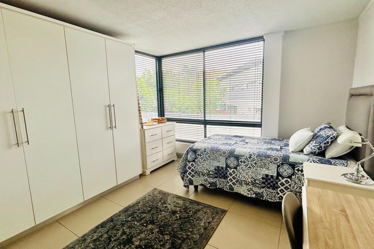 A bright bedroom featuring large corner windows with venetian blinds, tiled flooring, and ample built-in wardrobe storage.