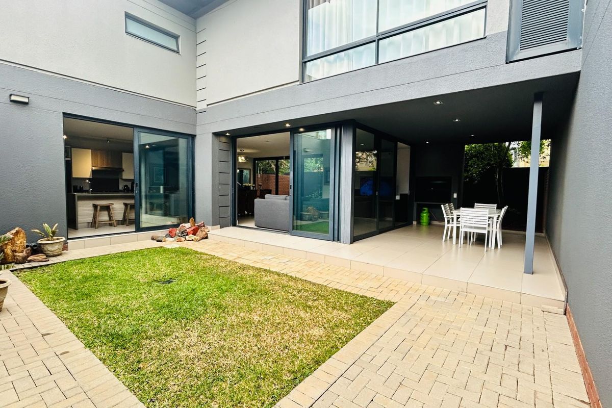 A modern rear courtyard featuring a covered patio area, paved flooring, and a small lawn, seamlessly connecting to the interior living spaces via sliding doors.