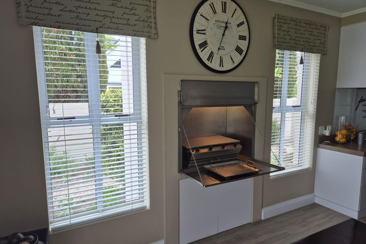 Indoor living area featuring a built-in stainless steel braai unit with integrated cabinetry and large windows with fitted blinds.