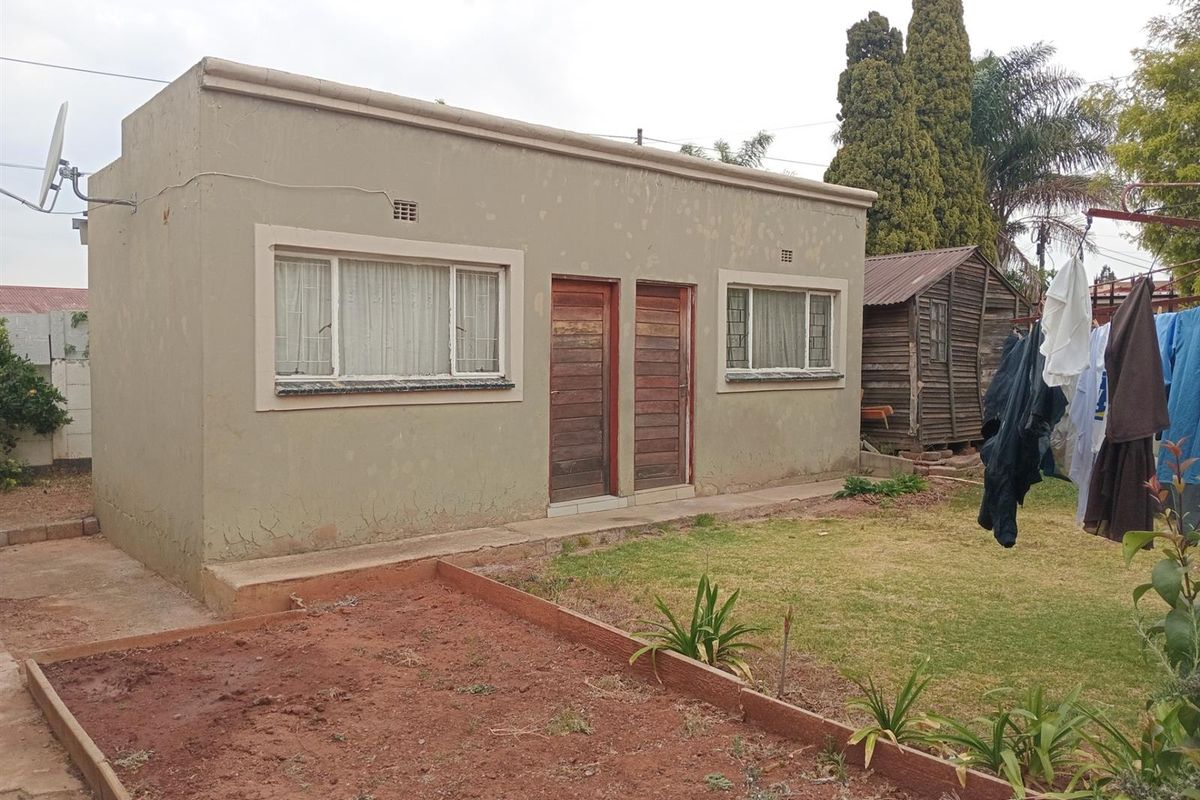 Rear view of a detached outbuilding featuring plastered masonry walls, timber doors, and steel-framed windows with security bars, set within a garden area.