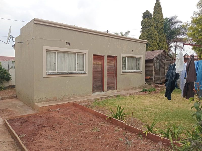 Rear view of a detached outbuilding featuring plastered masonry walls, timber doors, and steel-framed windows with security bars, set within a garden area.