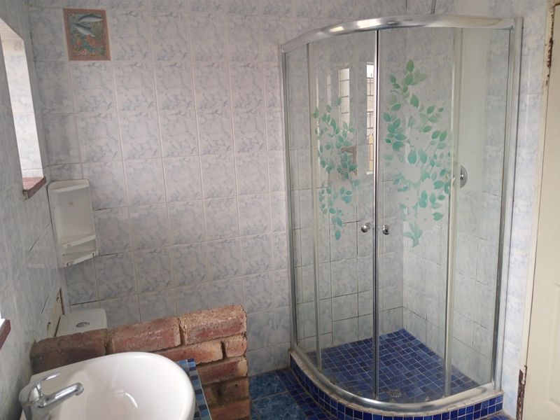 A dated bathroom featuring a curved glass shower enclosure, blue mosaic floor tiling, and a pedestal washbasin with exposed brick partition wall.