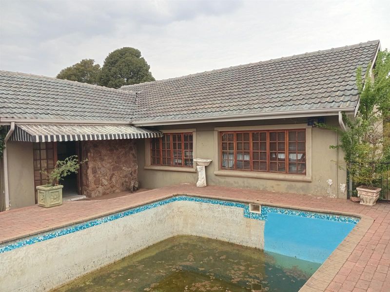 Rear exterior view featuring a swimming pool with brick patio, stone feature wall, and retractable awning, requiring significant maintenance and pool restoration.