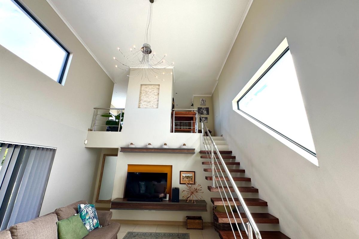 Spacious double-volume living area featuring a modern floating staircase, stainless steel balustrades, and a built-in media console with high ceilings and large windows.