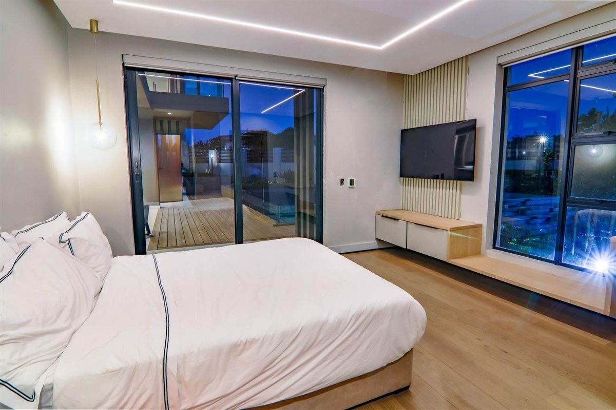 Modern bedroom featuring timber flooring, recessed LED ceiling lighting, built-in media console, and large sliding glass doors opening to an outdoor deck area.