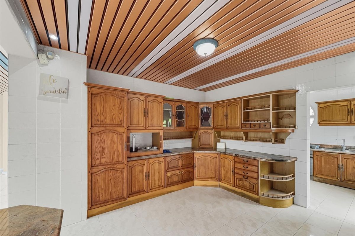 A traditional kitchen featuring extensive wooden cabinetry, tiled flooring, and a slatted ceiling, with an adjacent scullery area visible.