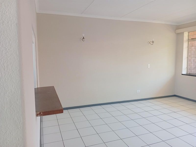 An empty, neutrally painted living area featuring white tiled flooring, wall-mounted lighting, and a convenient serving hatch connecting to the kitchen area.