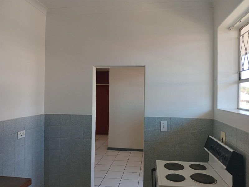 A functional kitchen space featuring a built-in electric stove, tiled splashbacks, and a window with security bars, leading into an adjacent room.