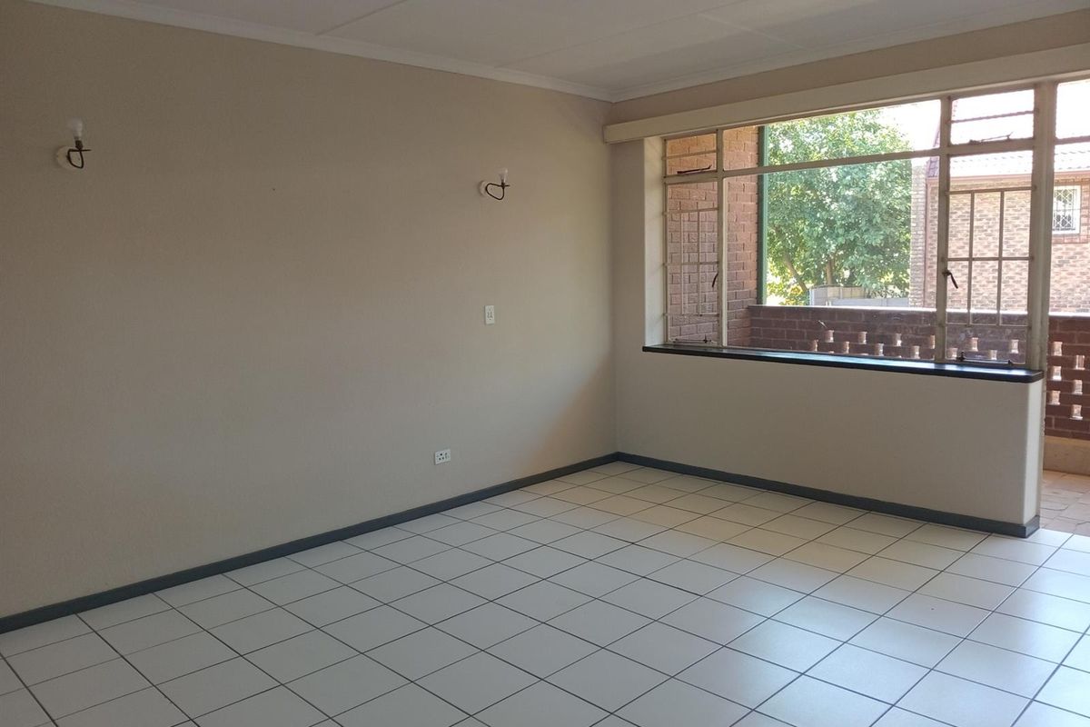 A bright, empty living room featuring neutral tiled flooring, large windows providing natural light, and wall-mounted lighting fixtures.